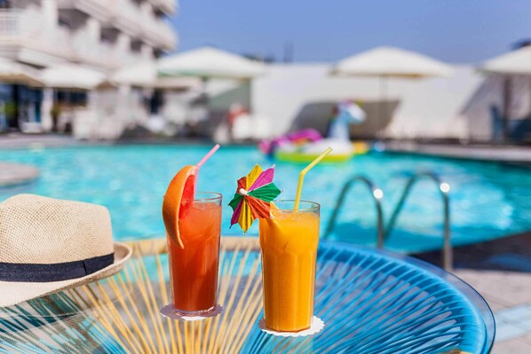 Une scène au bord de la piscine, avec deux boissons sur une table, un chapeau et une piscine en arrière-plan.