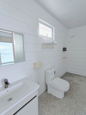Un baño moderno con lavamanos, inodoro y zona de ducha blancos. Cuenta con una ventana que ofrece luz natural y vistas al mar.
