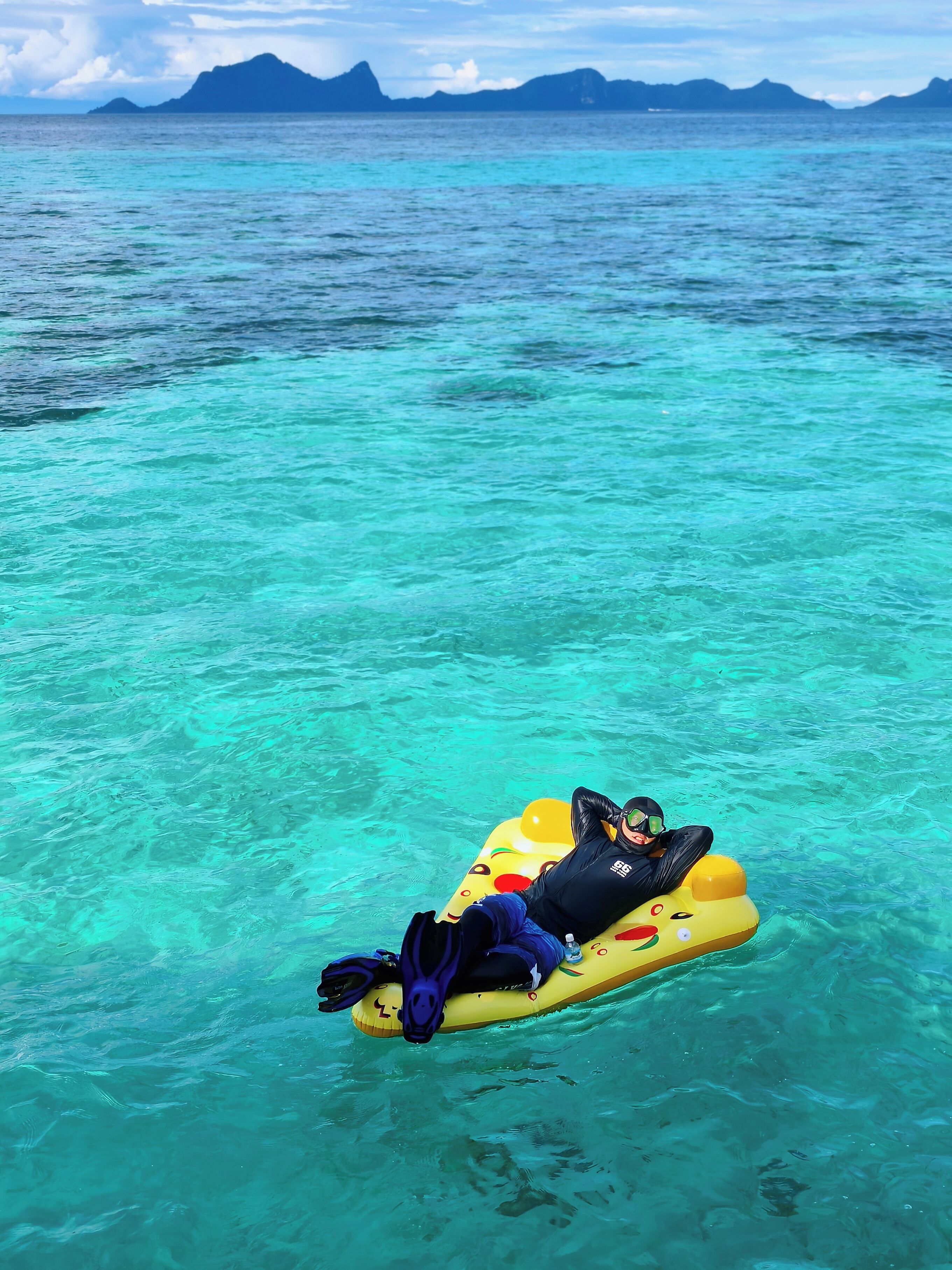 A person floating on a yellow inflatable raft in clear turquoise water with distant mountains.