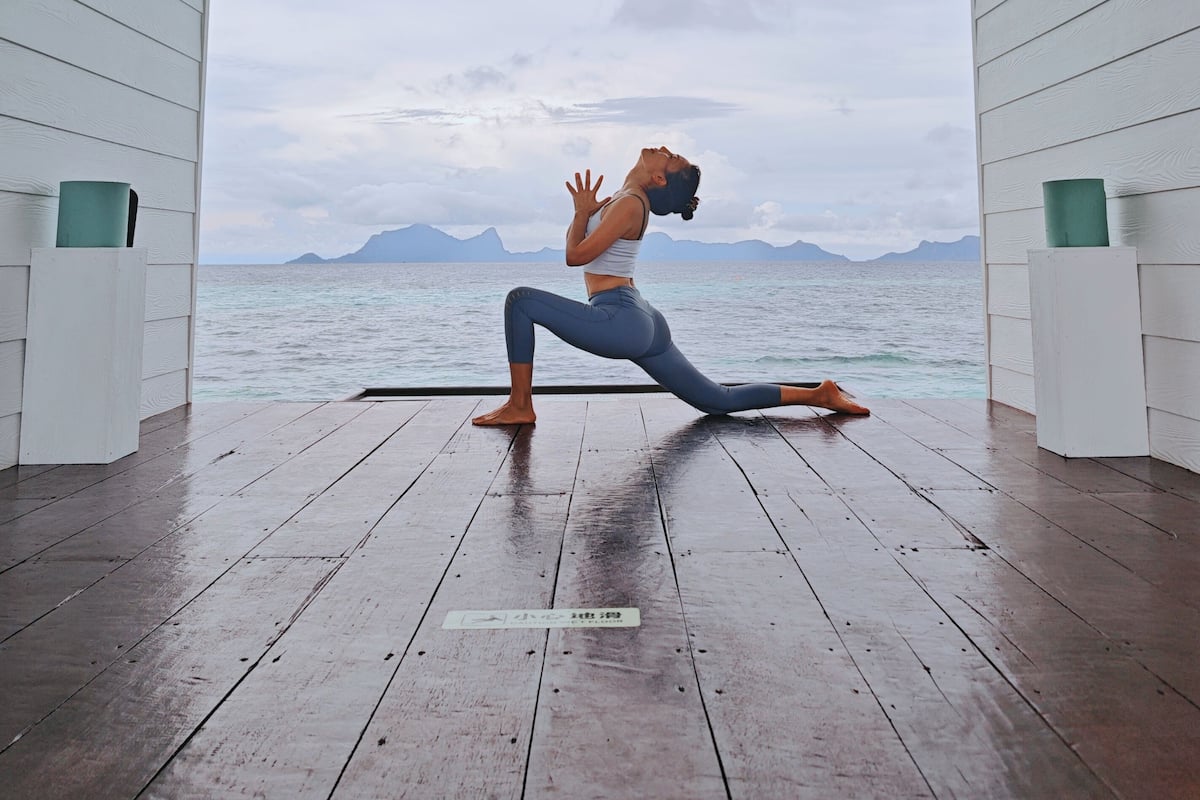 A person performing a yoga pose on a wooden deck with a view of the sea and mountains.