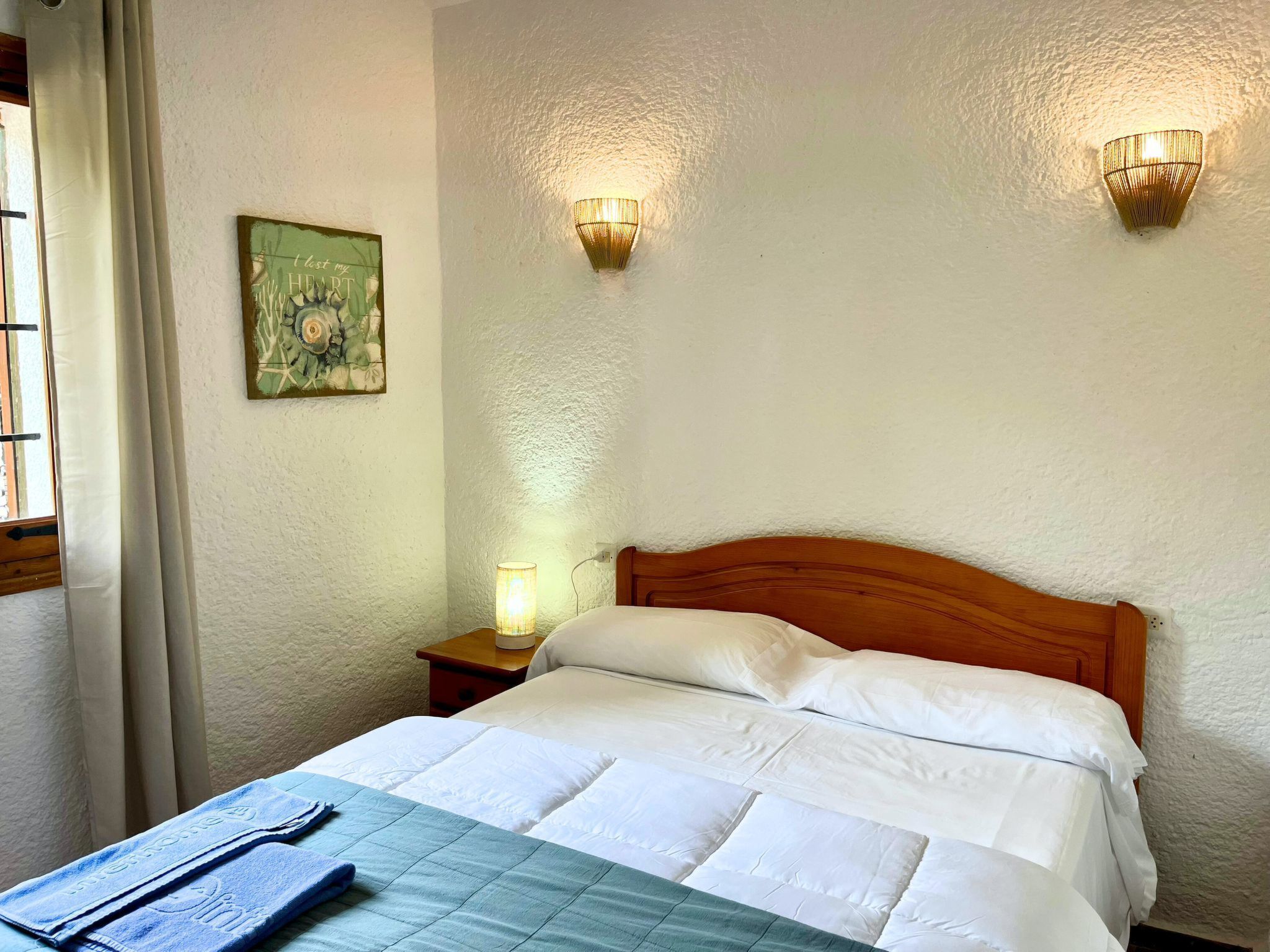 A neatly made bed with white linens, a blue quilt, and a blue towel on the side. There are two wall-mounted light fixtures and a small bedside table with a lamp. A framed picture hangs on the wall above the bed.