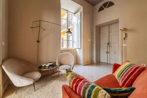 A room with a sofa, armchair, floor lamp, and a window with a view of buildings.