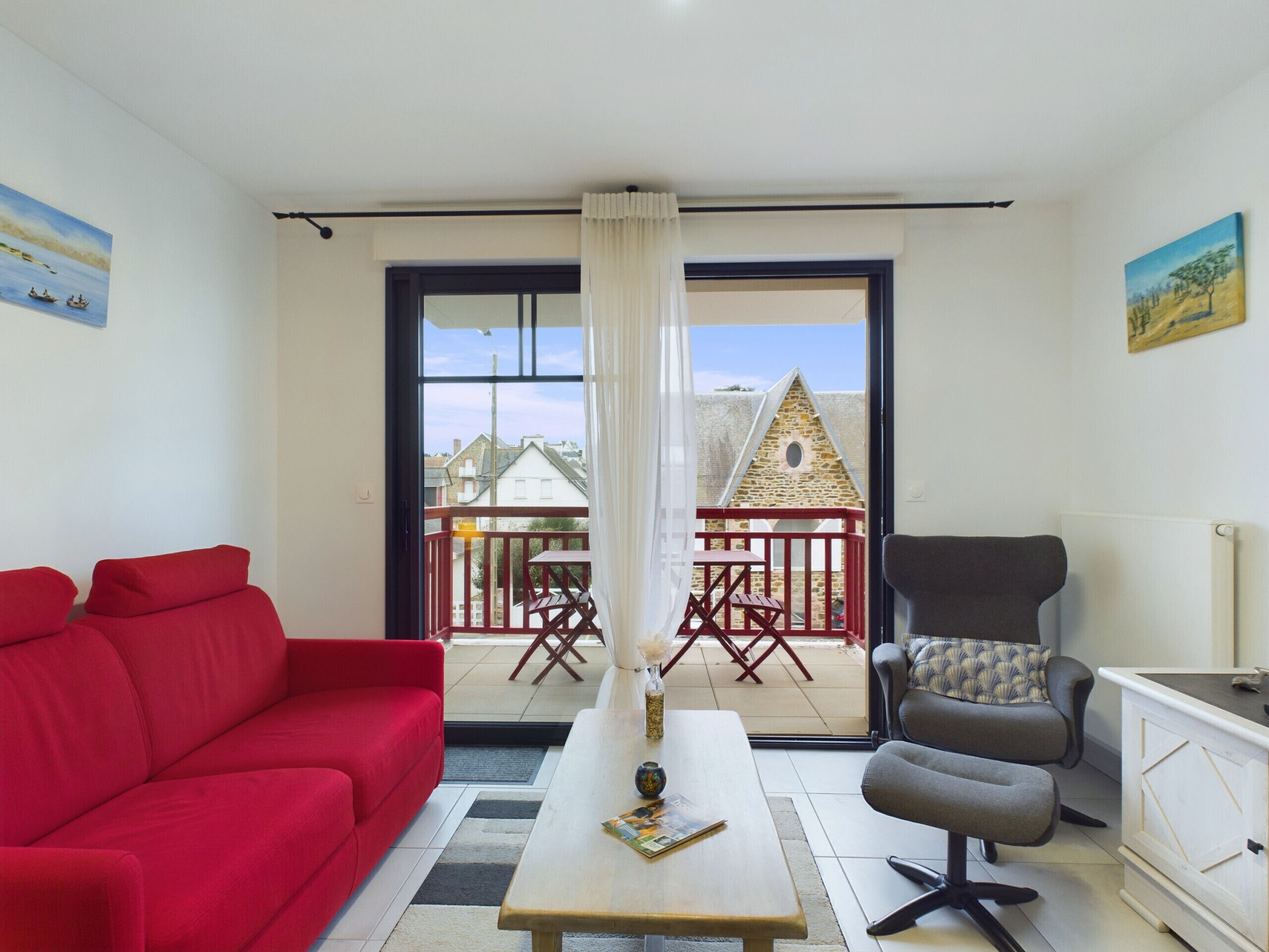 A living room with a red sofa, a grey armchair, a wooden coffee table, and a view of a balcony with a table and chairs.