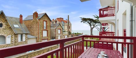 A balcony with a red railing overlooking stone buildings and a clear view of the sea.