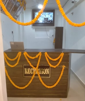 Mostrador de recepción con un letrero que dice 'RECEPTION', rodeado de guirnaldas de flores amarillas, una computadora portátil y una pantalla de televisión al fondo.