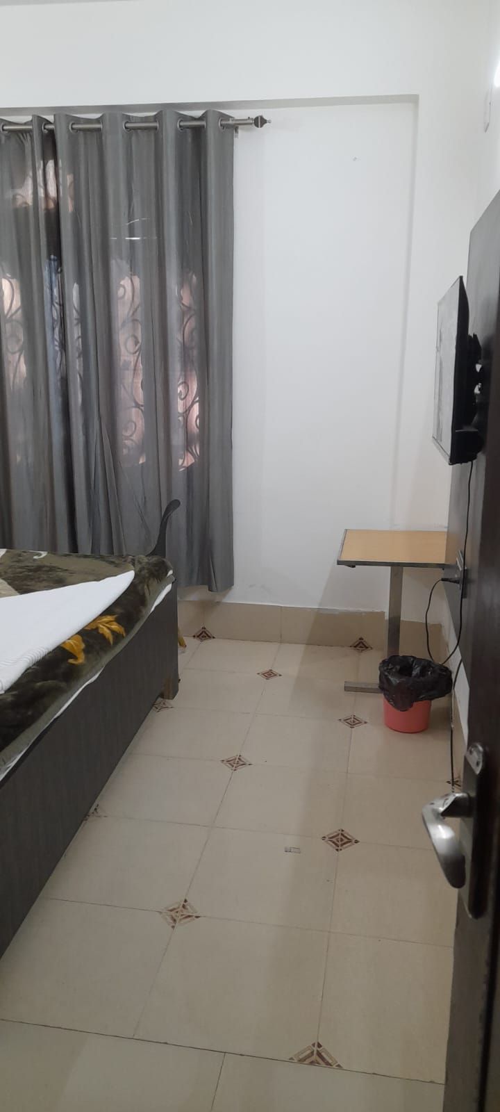 A room with a bed, a small table, a TV mounted on the wall, and a trash bin.
