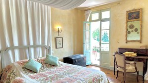 Een slaapkamer met een bed, een bureau en een stoel. Er is een raam met gordijnen en een deur naar buiten.
