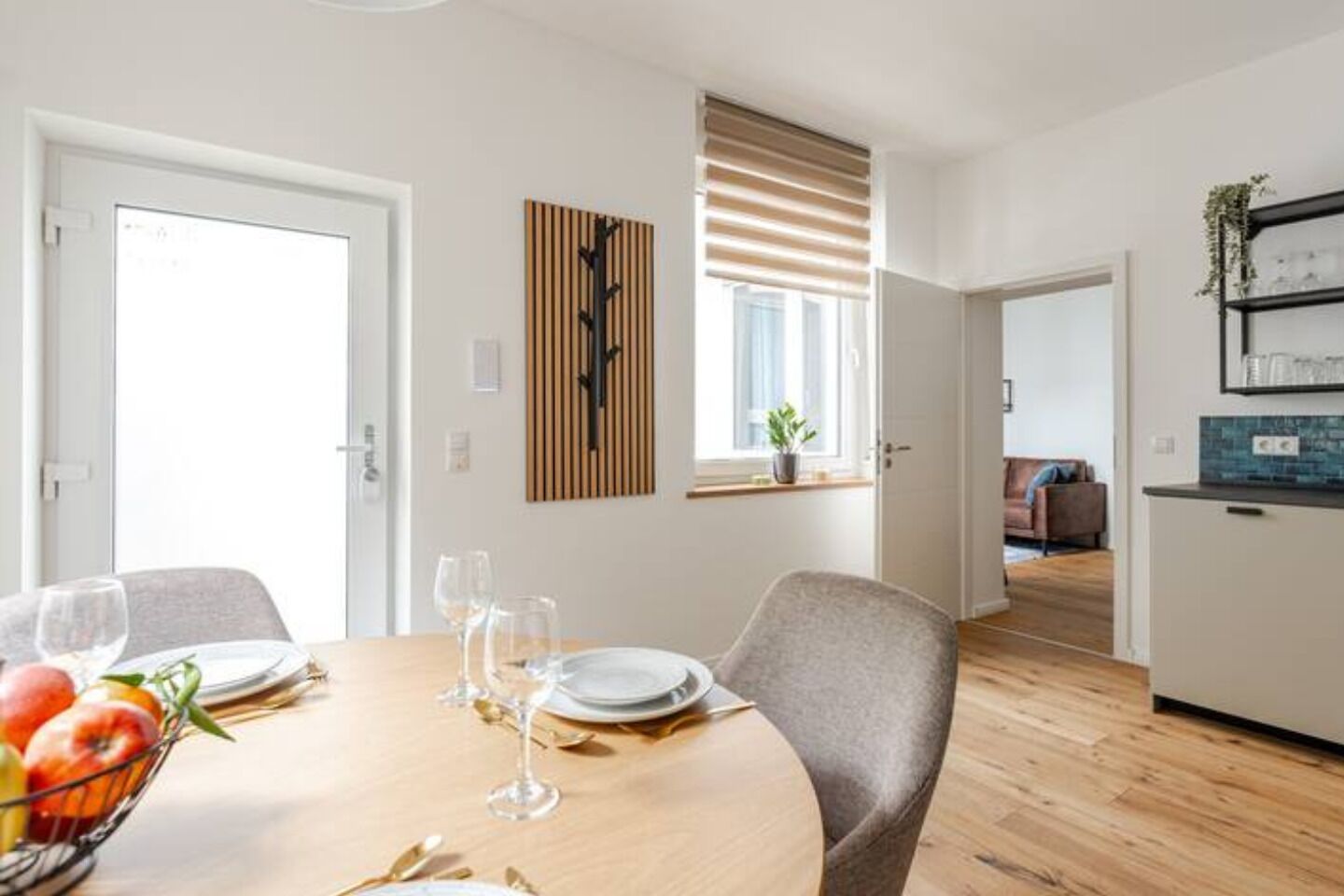 Un comedor moderno con una mesa de madera dispuesta para cuatro personas, una cocina al fondo y una ventana con persianas.