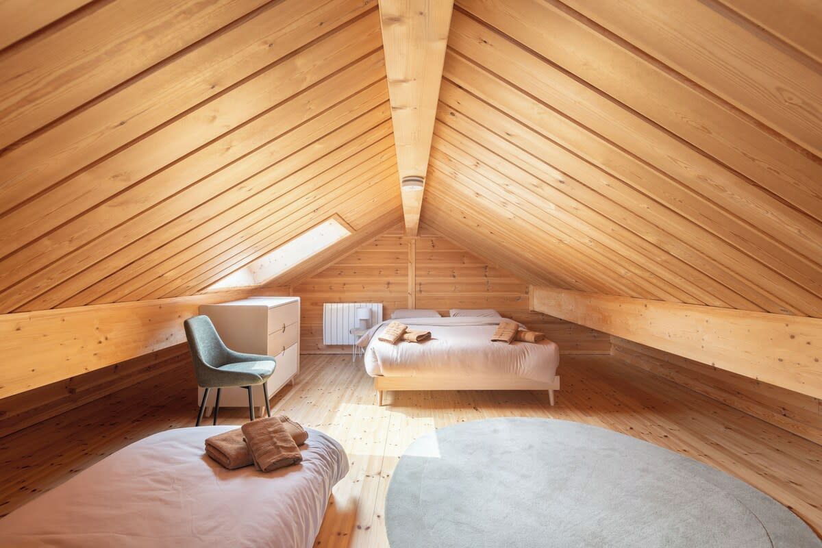 Une chambre mansardée avec un plafond haut et une charpente en bois inclinée, comprenant un lit, une chaise et un petit bureau.