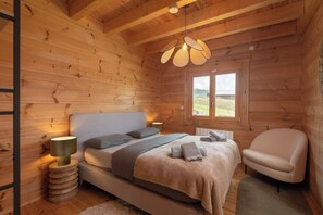 A wooden cabin bedroom with a bed, bedside lamps, a chair, and a window with a view. - Toison D'or - 8 pers. - Jacuzzi + sauna (Xonrupt-Longemer)