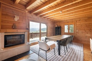 A wooden interior with a fireplace, dining table, chairs, and a view through large glass doors.