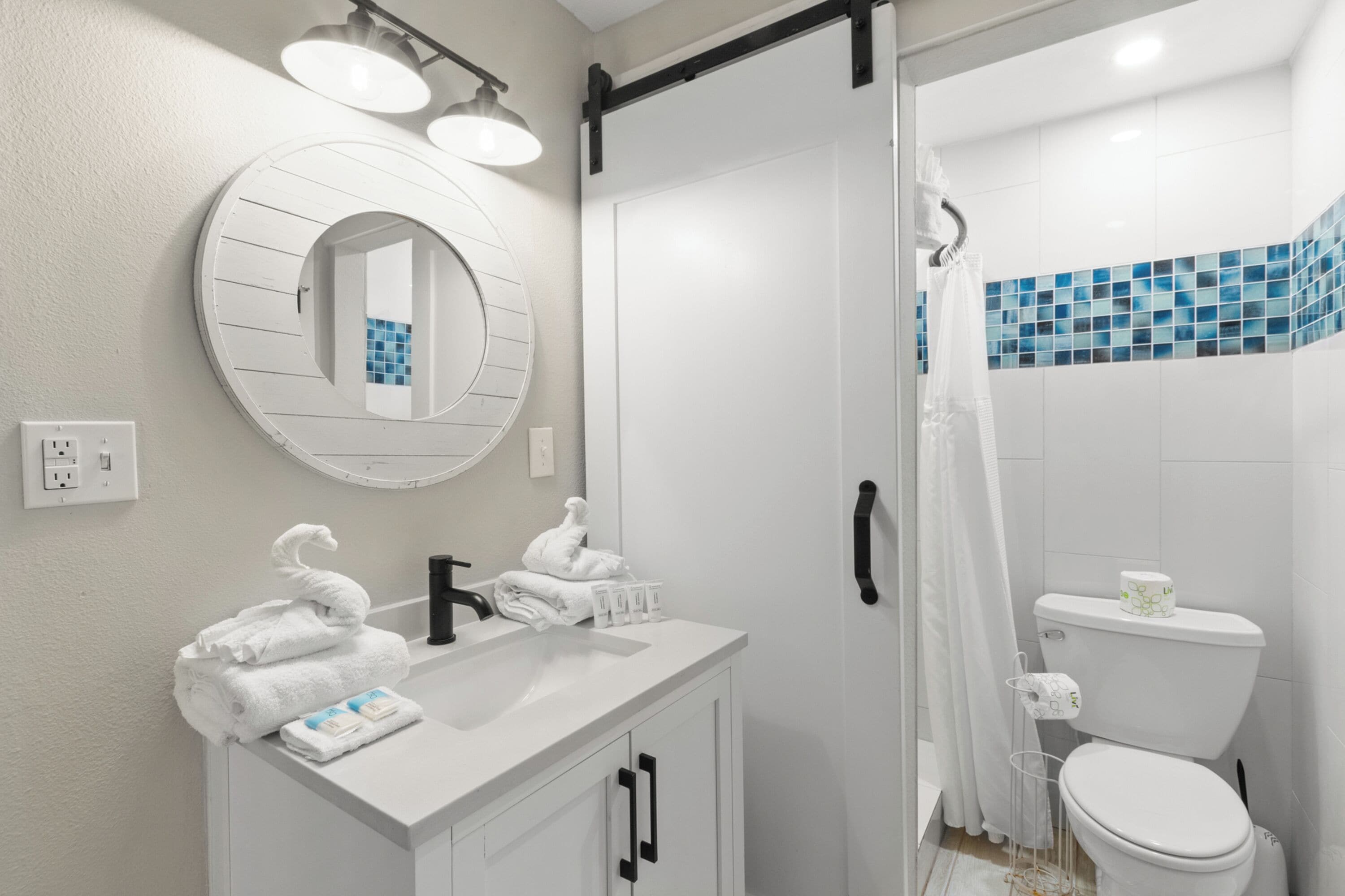 A modern bathroom with a white vanity, a round mirror, a shower with a glass door, and a toilet with a blue-tiled shower area.