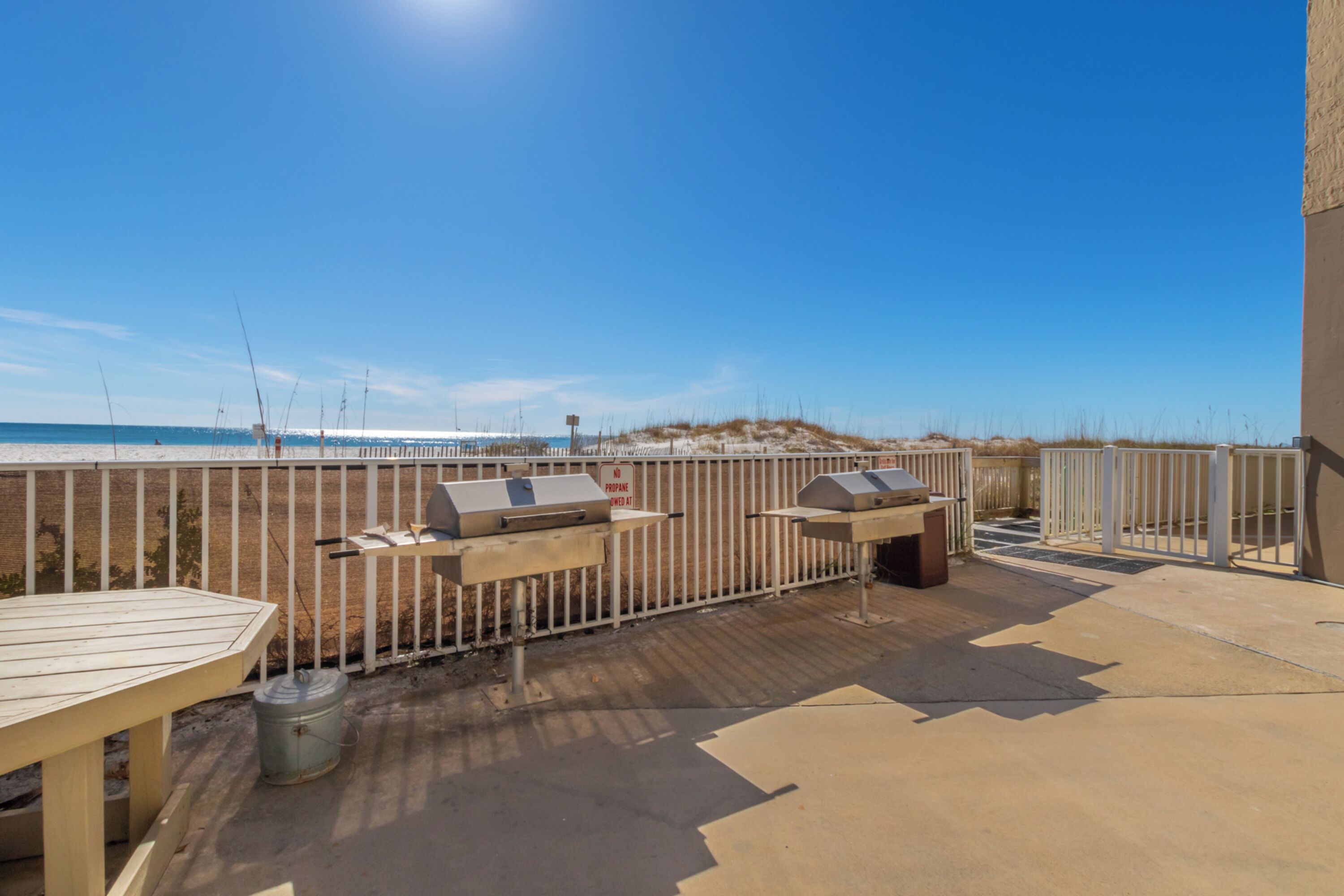 A patio with a grill and a table, overlooking a beach with a fence and dunes.