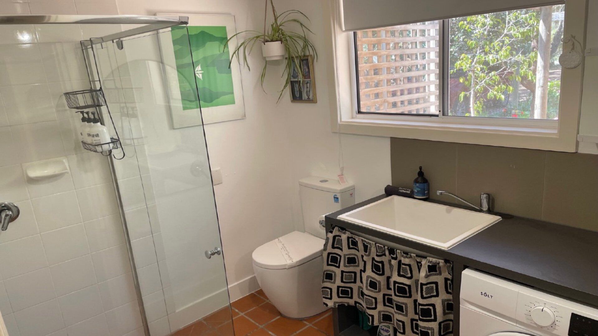 Een badkamer met een glazen douchecabine, toilet, wastafel met een handdoek met dessin, en een raam met uitzicht op bomen.