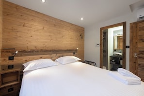 Ein Hotelzimmer mit einem hölzernen Kopfteil, einem Bett mit weißen Leinen, einem Nachttisch mit einer Lampe und einem durch eine offene Tür einsehbaren Badezimmer.