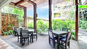 An outdoor patio with tables and chairs, surrounded by lush greenery and a view of a residential area.