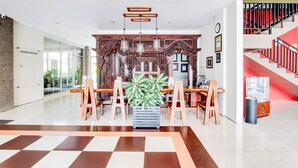 A spacious lobby with a checkered floor, a central plant, wooden chairs, and a staircase with red railings.