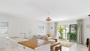 Una cocina moderna con armarios blancos, encimera de madera y un fregadero. Hay una zona de comedor con una mesa y sillas, y un sofá al fondo.