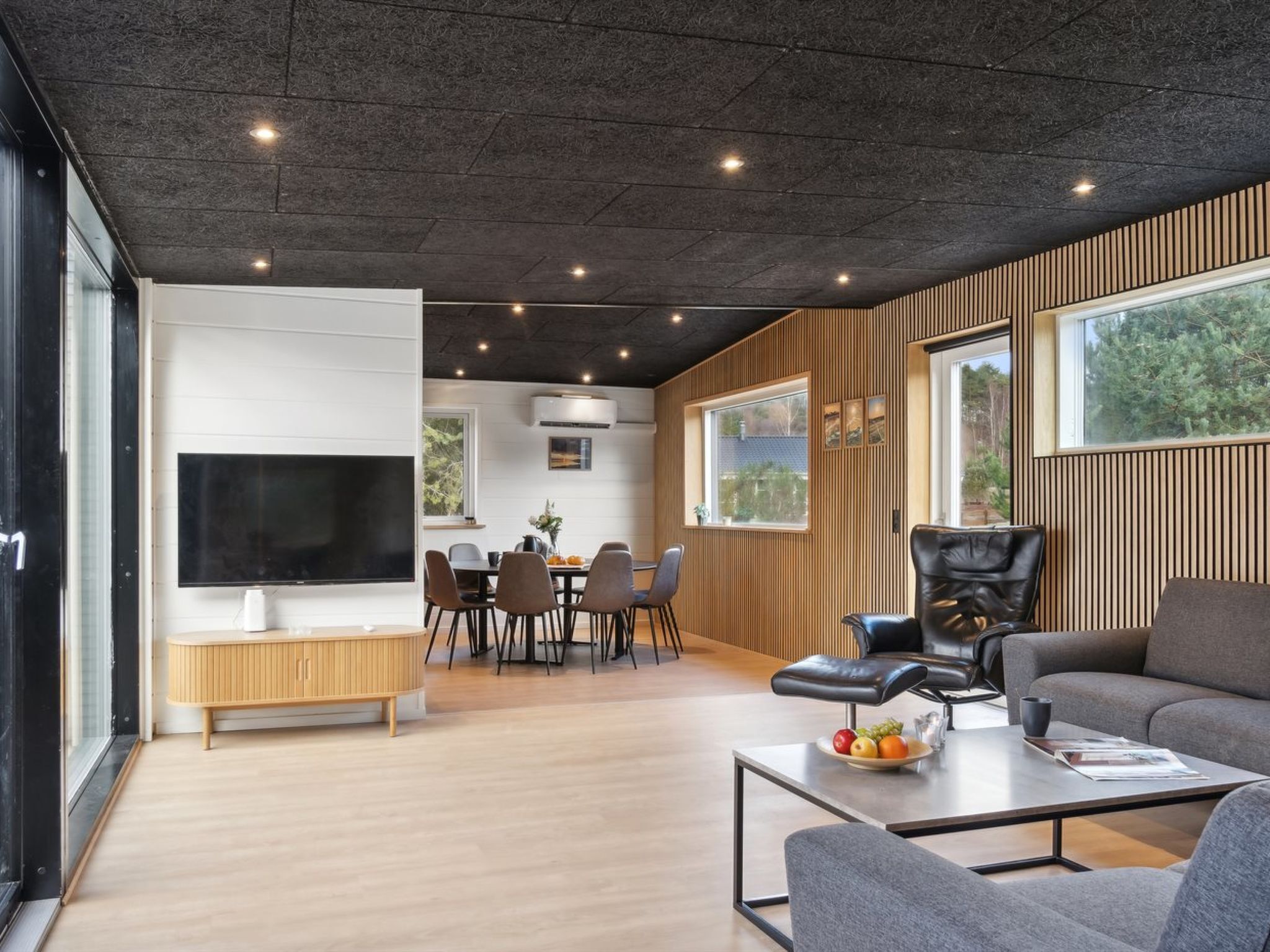 A modern living room with a flat-screen TV, a dining area with a wooden table and chairs, and a seating area with a leather armchair and sofa.