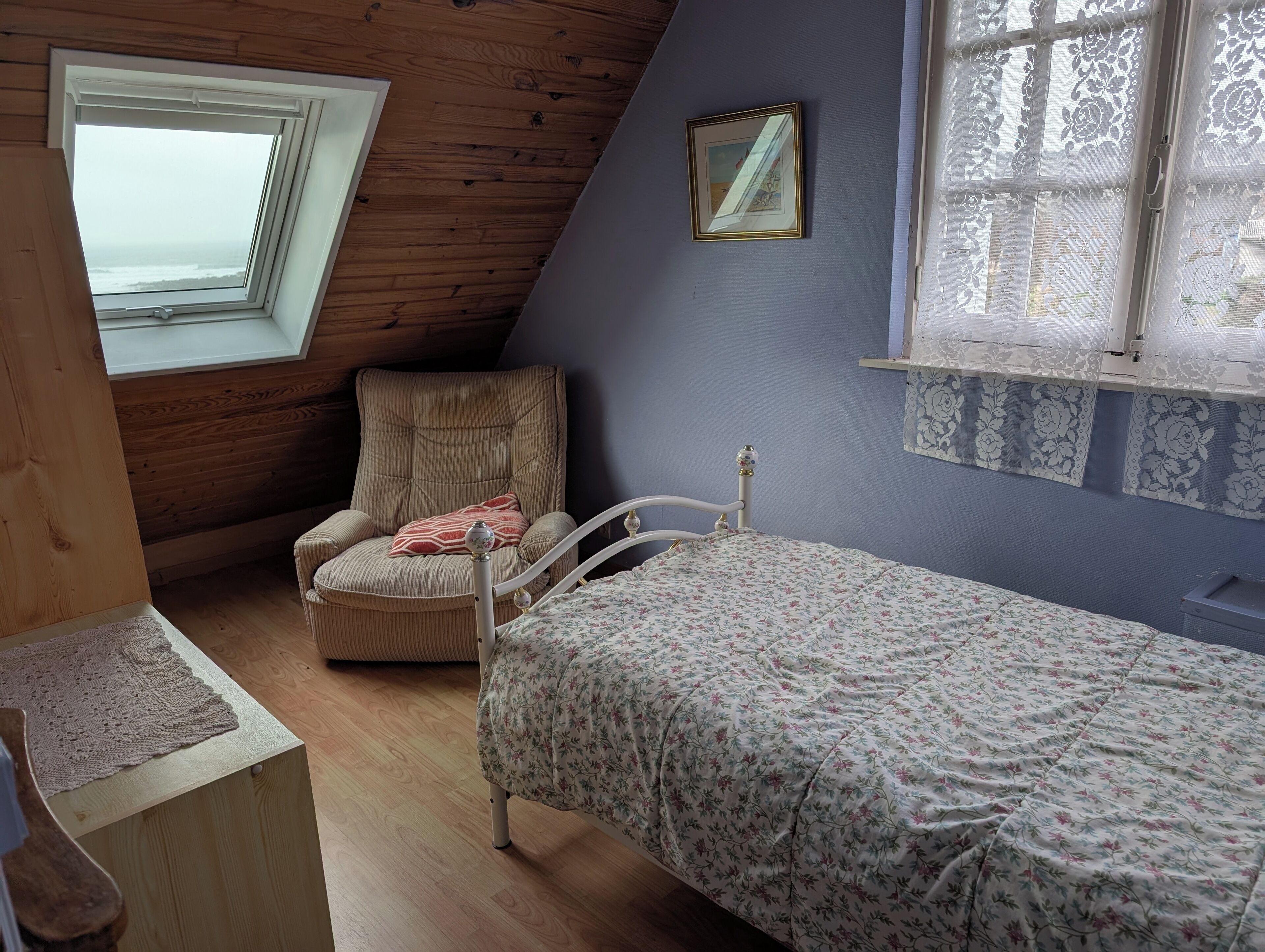 A room with a sloped ceiling, a single bed with a floral bedspread, a cushioned chair, a small table, and a window with a view of the ocean.