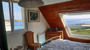 Un dormitorio con cama, una silla de mimbre, un aparador con una radio y un cuadro.