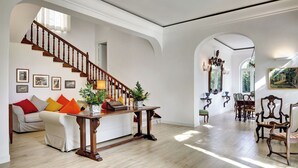 A spacious living room with a wooden staircase, a dining area with antique furniture, and a view of the outdoors through large windows.