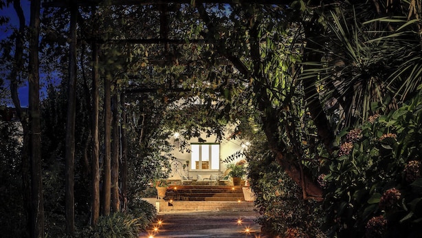 A pathway leading to a building framed by a wooden archway, surrounded by lush greenery and illuminated by small lights.