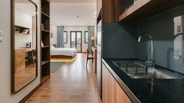 A modern hotel room with a large mirror, wooden furniture, a bed, a desk, and a kitchen area with a sink and stove.
