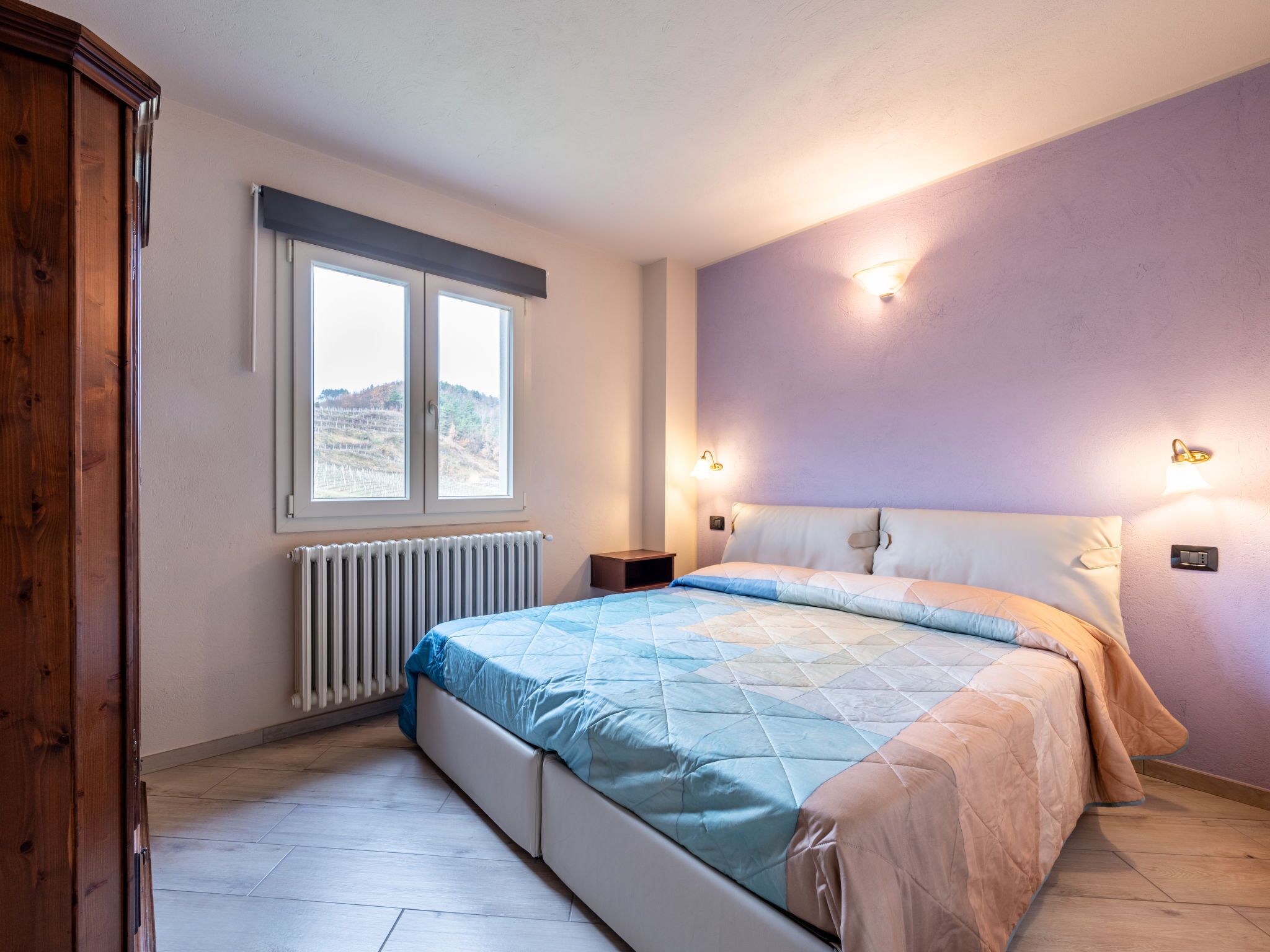 A bedroom with a large bed, a window with a view, a radiator, and wall-mounted lights.