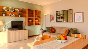 A living room with a TV, wooden cabinets, a sofa, and a dining table with a fruit bowl. - Apartment Paníco by Interhome (Dorio)