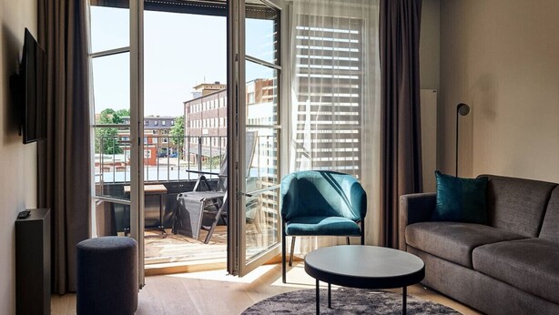 Ein modernes Wohnzimmer mit einer Couch, einem kleinen runden Tisch und einem blauen Sessel. Ein großes Fenster mit Jalousien, und ein Balkon ist von außen sichtbar.