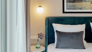 Ein Hotelzimmer mit Bett, Nachttisch mit Vase, Wandlampe und einem gerahmten Stadtbild.