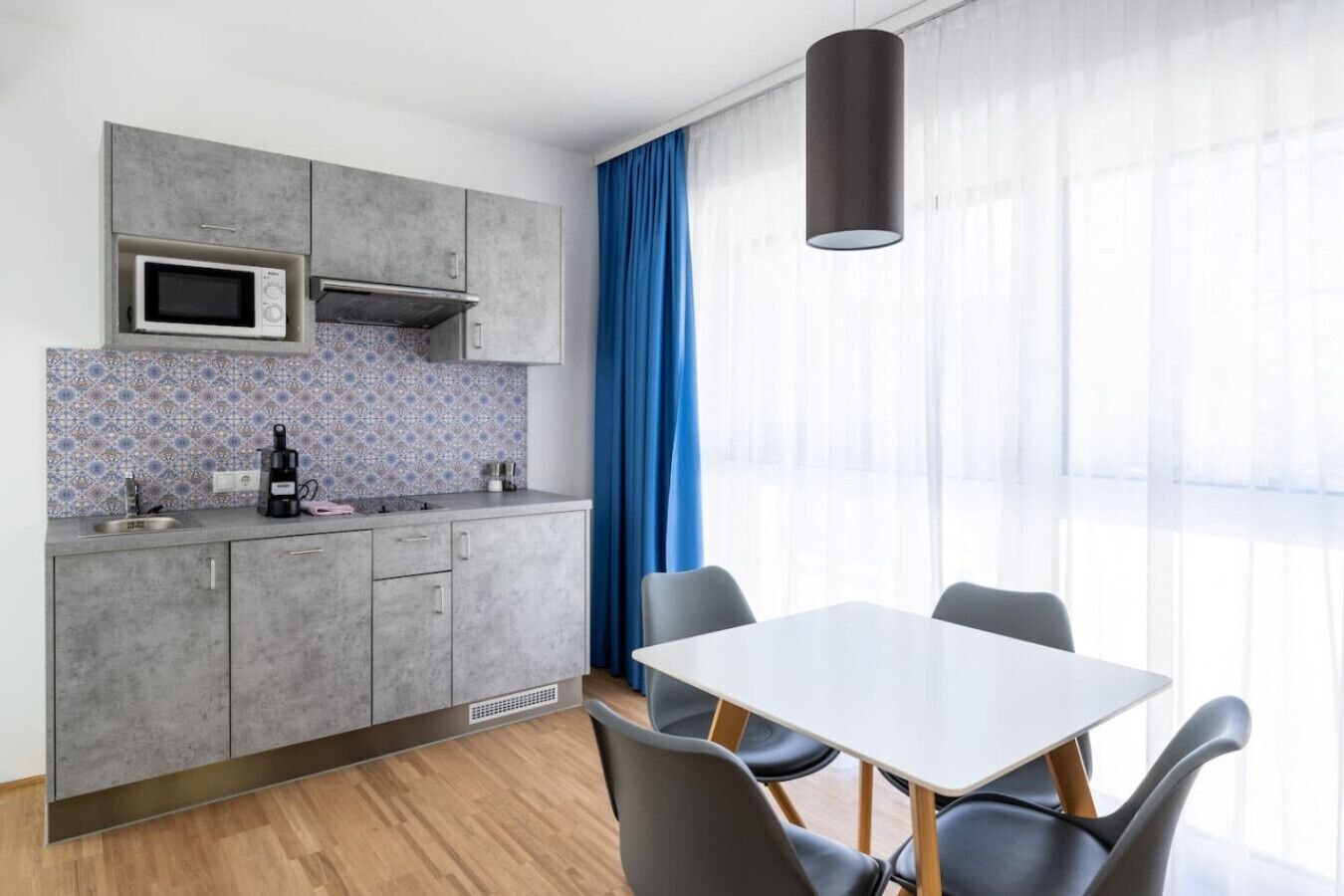Eine moderne Küche mit grauen Schränken, einer Mikrowelle und einer Kaffeemaschine. Ein Esstisch mit Stühlen und ein großes Fenster mit blauen Vorhängen sind ebenfalls vorhanden.