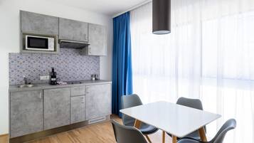 Eine moderne Küche mit grauen Schränken, einer Mikrowelle und einer Kaffeemaschine. Ein Esstisch mit Stühlen und ein großes Fenster mit blauen Vorhängen sind ebenfalls vorhanden.
