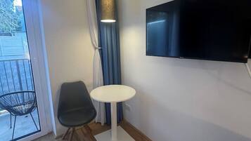 A modern room with a wooden floor, a round white table, two chairs, a TV mounted on the wall, and a window with curtains.