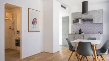 A modern kitchen with grey cabinets, a microwave, and a sink. There is a dining area with a white table and grey chairs. A framed bird painting hangs on the wall.