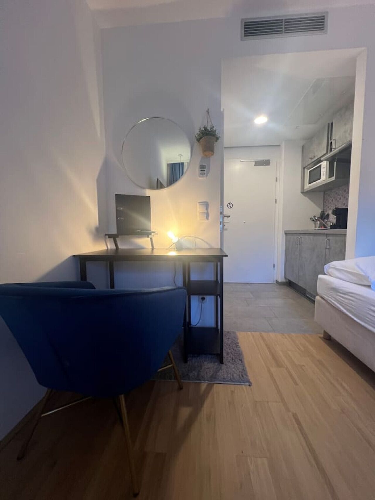 A compact living space with a desk, chair, and a bed. A small kitchen area is visible in the background.