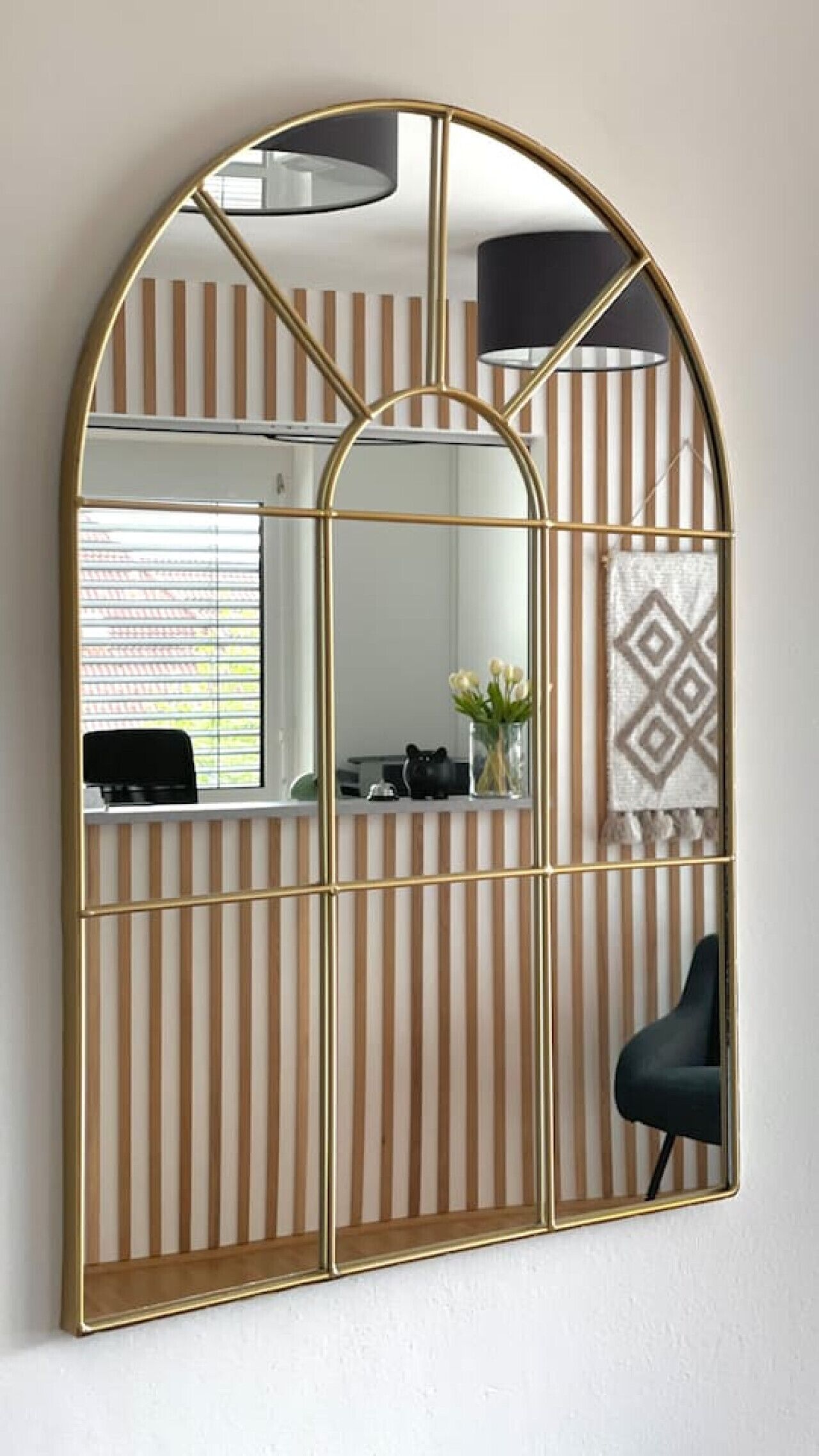 A room with a large arched mirror, a black chair, a desk with a computer, and a window with blinds.