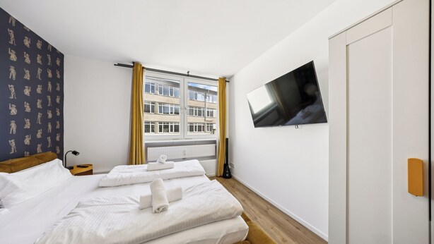 A hotel room with a bed, a TV, a window with curtains, and a view of buildings outside. - Arbio I Cube Apartments (Hamburg)