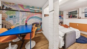 A room with a colorful brick wall mural, a blue table with orange chairs, a bed with a wooden headboard, and a window with a pink flamingo decoration. - Arbio I Cube Apartments (Hamburg)