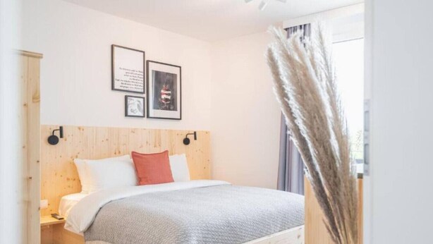 Una camera da letto con una testiera in legno, un letto con biancheria bianca e grigia, un cuscino rosso e due quadri appesi al muro.