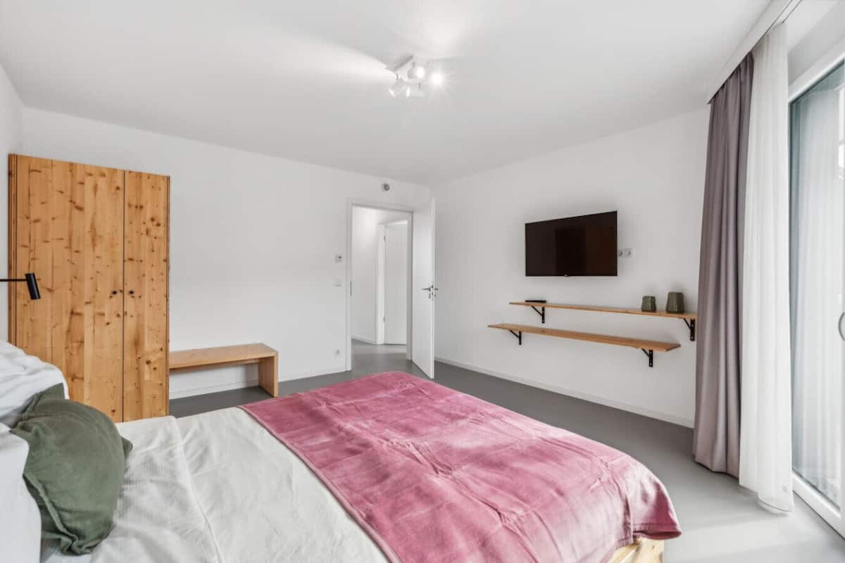 Ein modernes Schlafzimmer mit Bett, einem Holzschrank, einer Bank, einem an der Wand montierten Fernseher und einem Fenster mit Vorhängen.