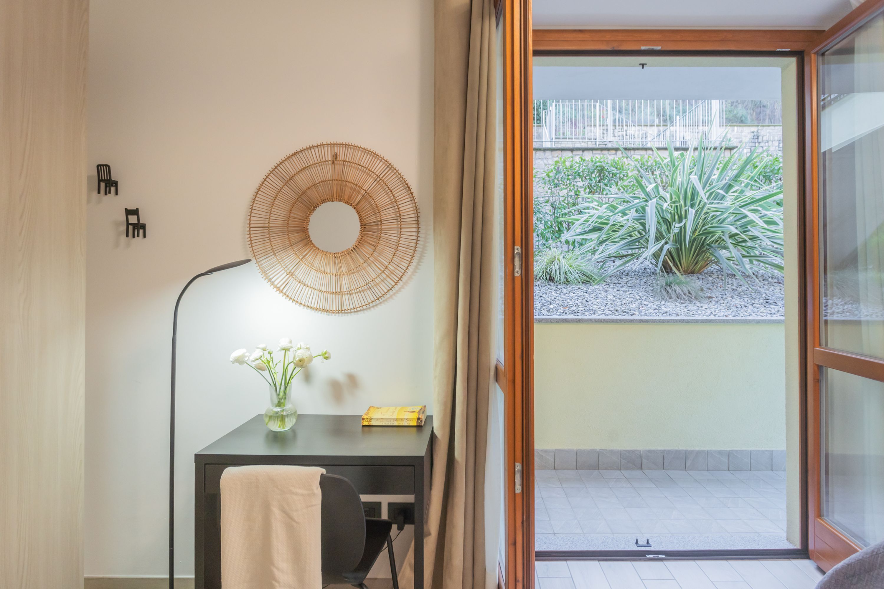Una habitación con escritorio, una silla, un florero con flores, una lámpara y vistas a un jardín por un ventanal corredera.