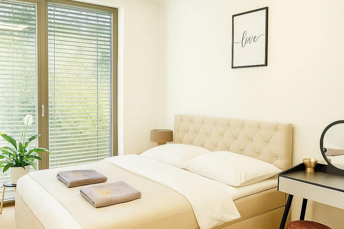 Een moderne slaapkamer met een groot bed, een nachtkastje, een klein krukje en een raam met jaloezieën.