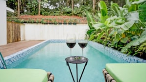 Une vue sur la piscine, avec deux chaises longues, une table sur laquelle reposent deux verres de vin rouge, et une végétation luxuriante en arrière-plan.