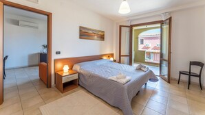 A bedroom with a bed, bedside lamps, a nightstand, a chair, and a view through an open door. - Le Fontane Apartments In Villasimius (Villasimius)