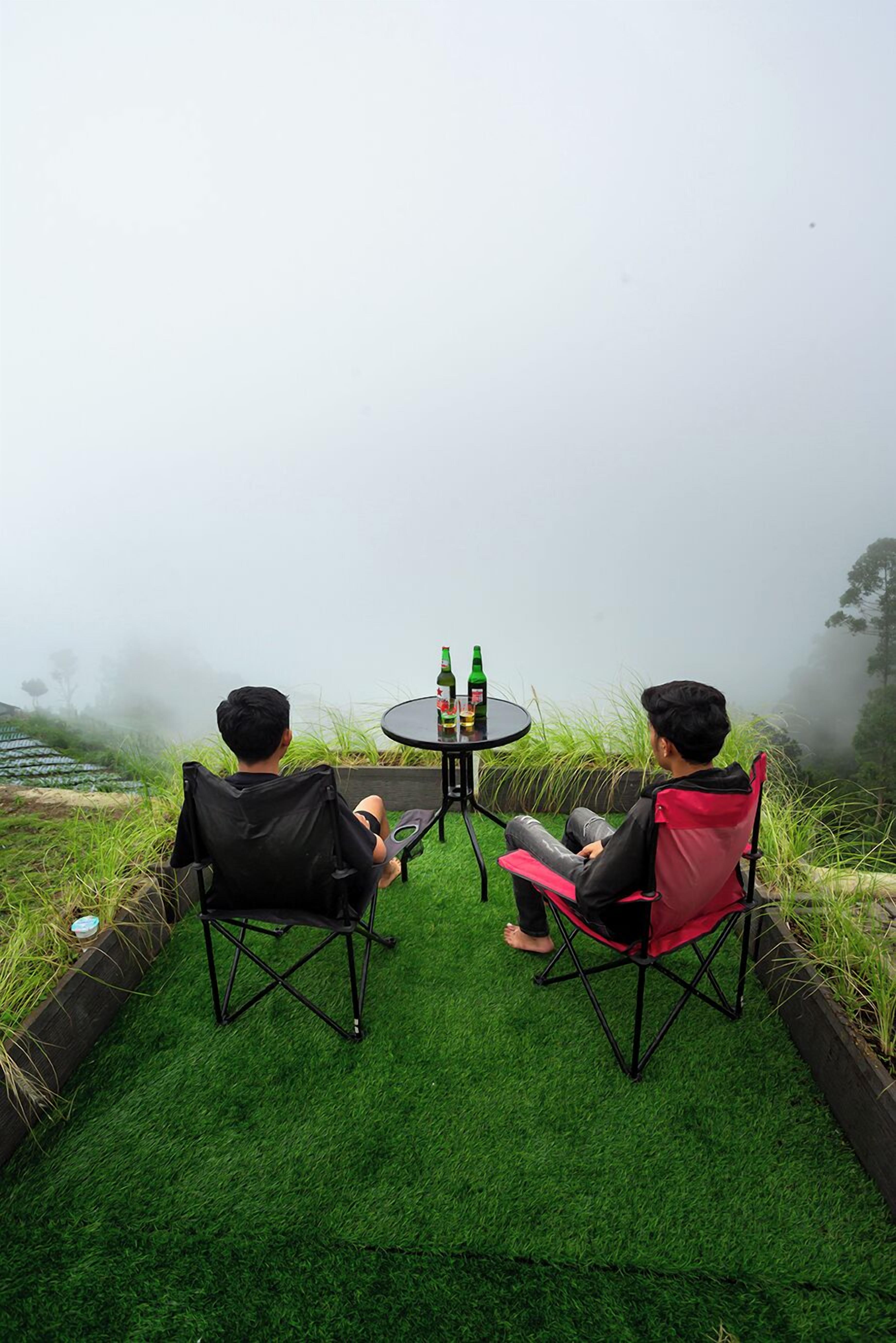 Two people sitting on folding chairs facing a table with bottles, surrounded by artificial grass and a foggy landscape.