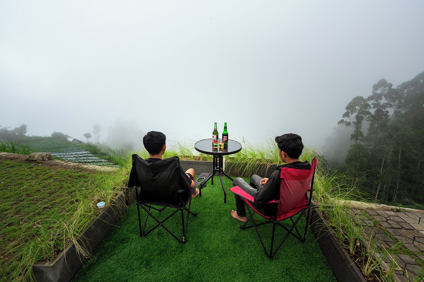 Two people sitting at a small round table with drinks, surrounded by artificial grass and a foggy landscape.