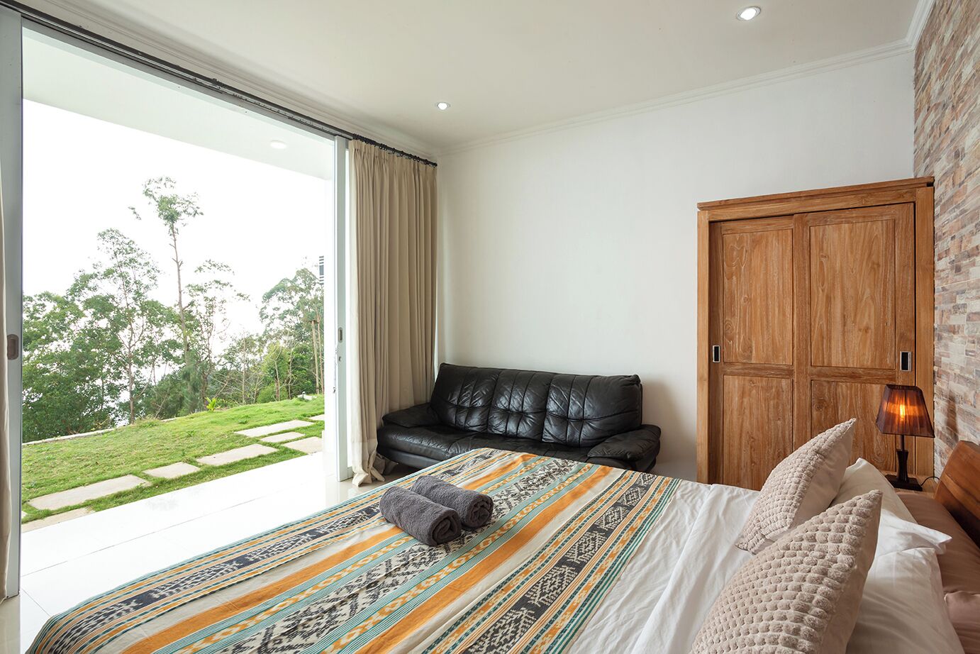 A bedroom with a large bed, a leather sofa, a wooden wardrobe, and a view of a garden.