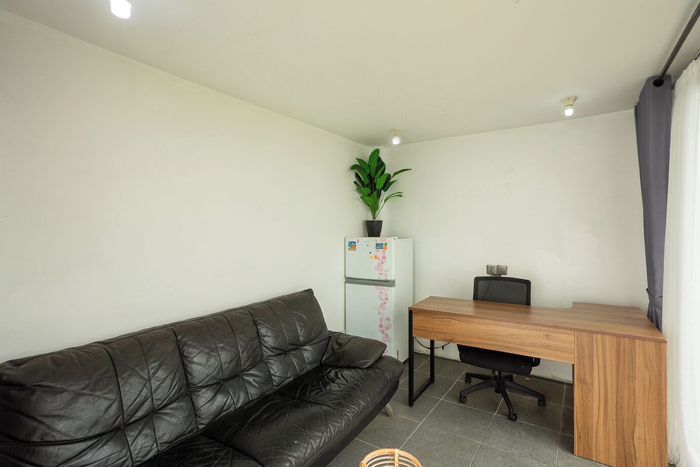 A compact living space with a black leather sofa, a wooden desk, and a white refrigerator with magnets.